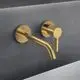 Wandarmatur in gebürstetem Gold von Ideal Standard mit runder Rosette und einem langen, schwenkbaren Auslauf. Montage an einer grauen Wandoberfläche.